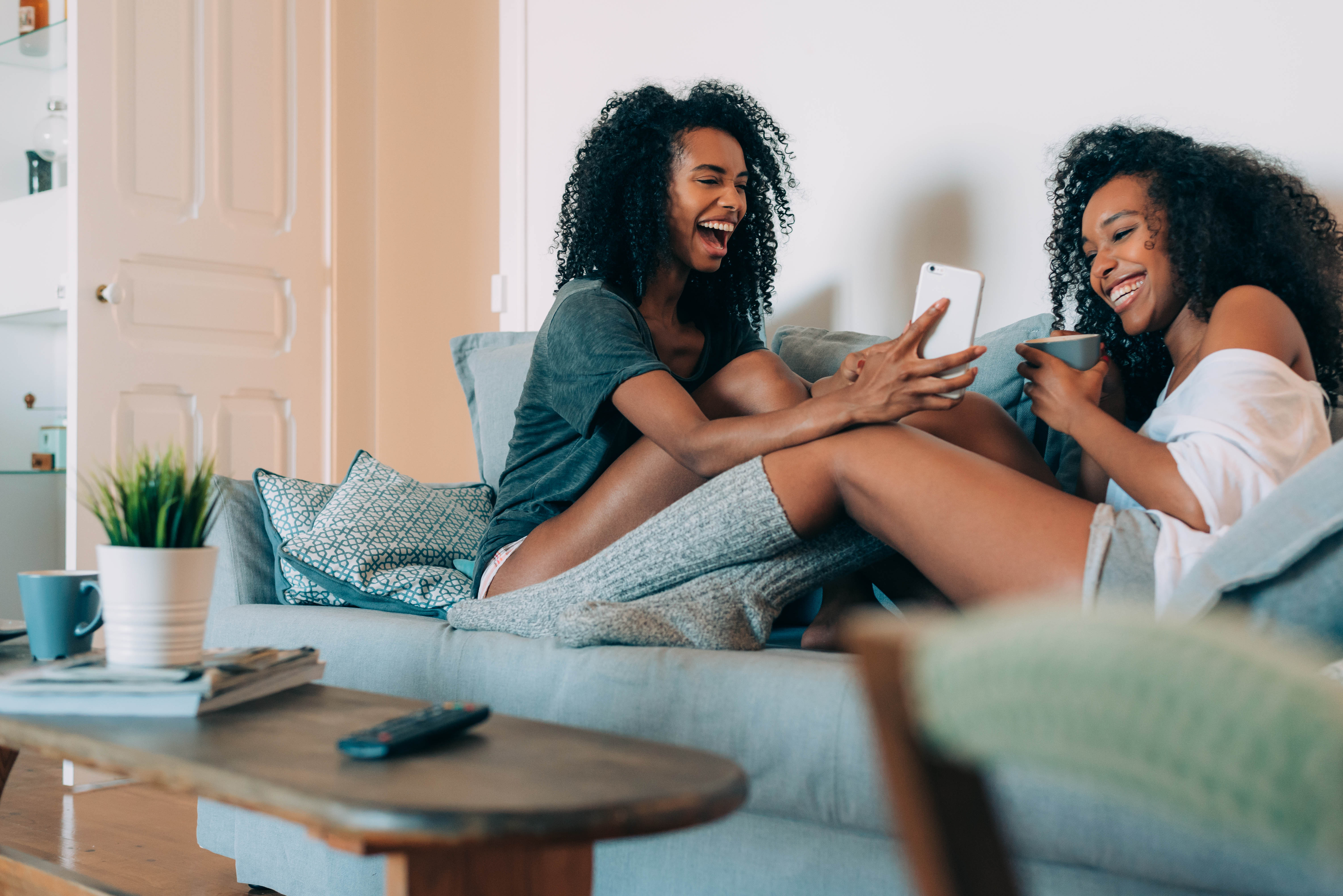 Two young black women sitting on a couch with their phones, laughing and talking